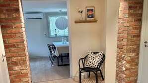 Une pièce avec un mur en briques, un coin repas avec une table et des chaises, et une vue sur l’extérieur par une porte ouverte.