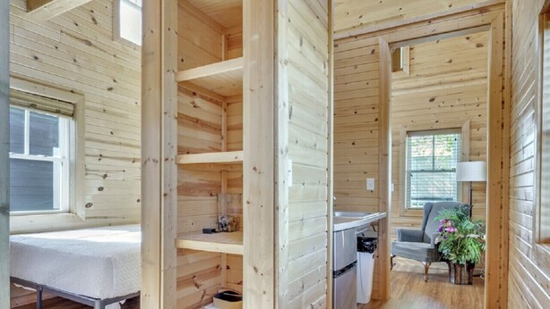A wooden cabin interior with a bed, shelves, a kitchen area, and a dining table.