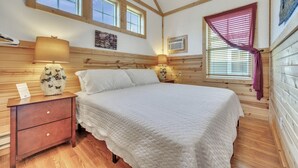 Un dormitorio con cama, cabecera de madera, mesitas de noche, lámparas, una ventana con persianas y un cuadro en la pared.