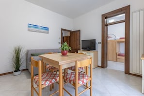 Ein Essbereich mit Holztisch und -stühlen, einer Topfpflanze, einem Fernseher und Blick auf ein Schlafzimmer mit Etagenbett.