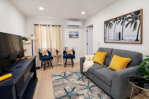 A living room with a grey sofa, two blue armchairs, a TV, and a painting of palm trees. - New! Modern 1BR Patio Apartment 3 blocks to Ocean (San Diego)