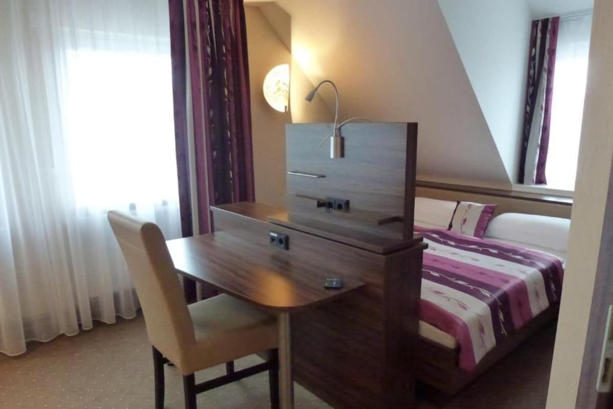 A hotel room with a bed, a desk with a chair, a bedside table, and a window with curtains.