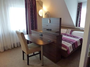 A hotel room with a bed, a desk with a chair, a bedside table, and a window with curtains.