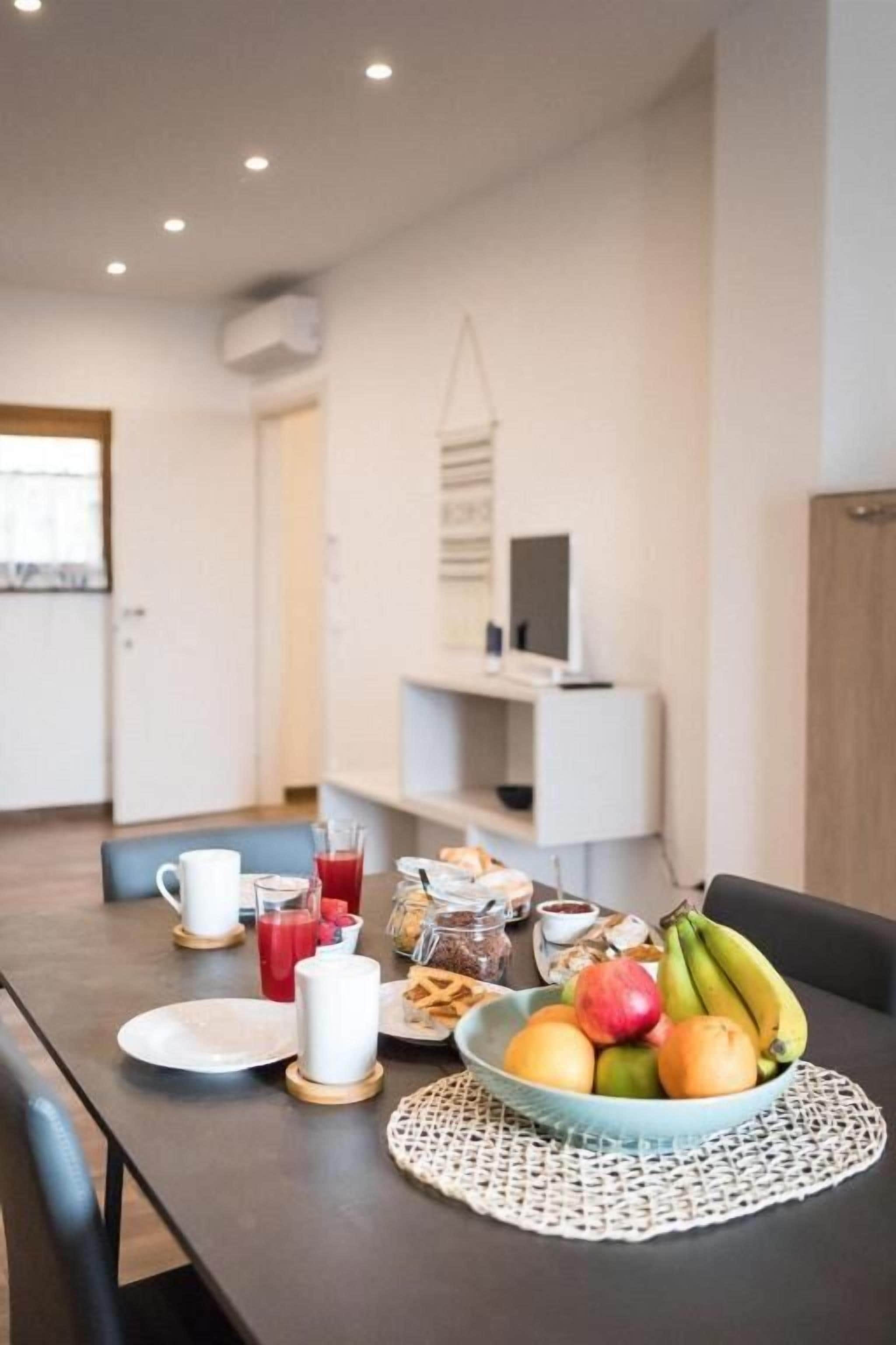 A dining area with a table set for breakfast, including fruits, bread, and beverages.
