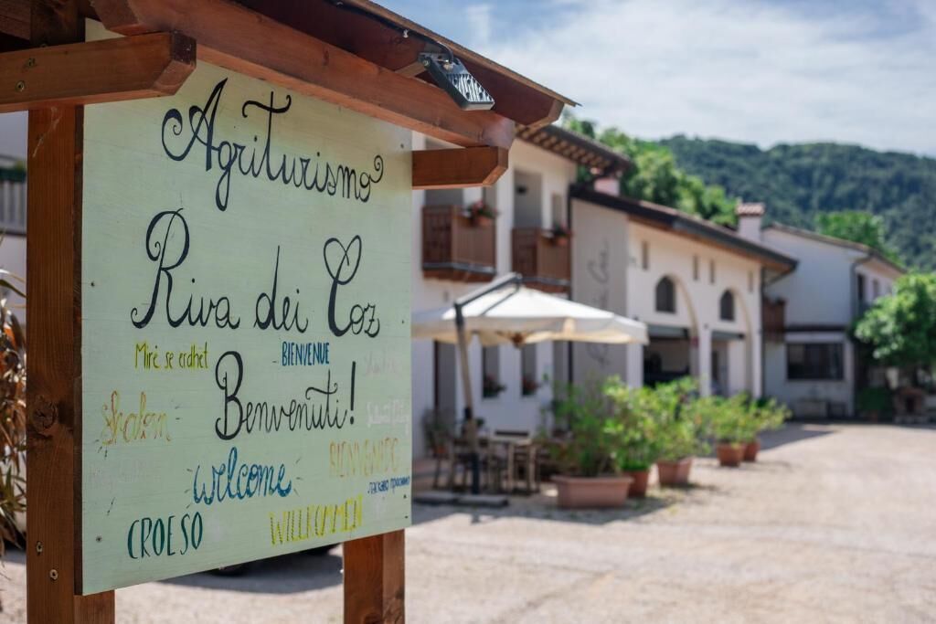 A signboard with multiple welcome messages in different languages, a wooden pergola, and a building with outdoor seating in the background.