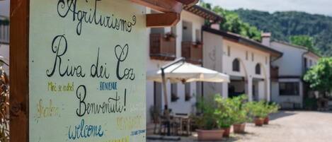 A signboard with multiple welcome messages in different languages, a wooden pergola, and a building with outdoor seating in the background.