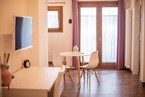 Ett rum med ett vitt bord, två stolar, en klocka, en tv på väggen och ett fönster med gardiner.