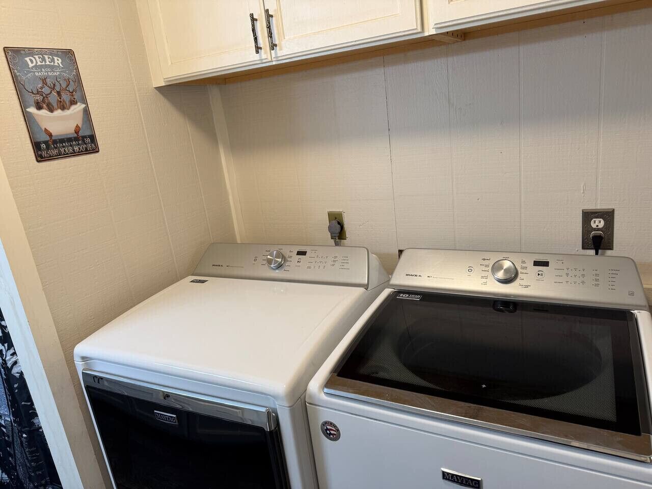 Een wasruimte met twee gestapelde wasmachines en drogers, een poster aan de muur, en een witte betegelde muur.