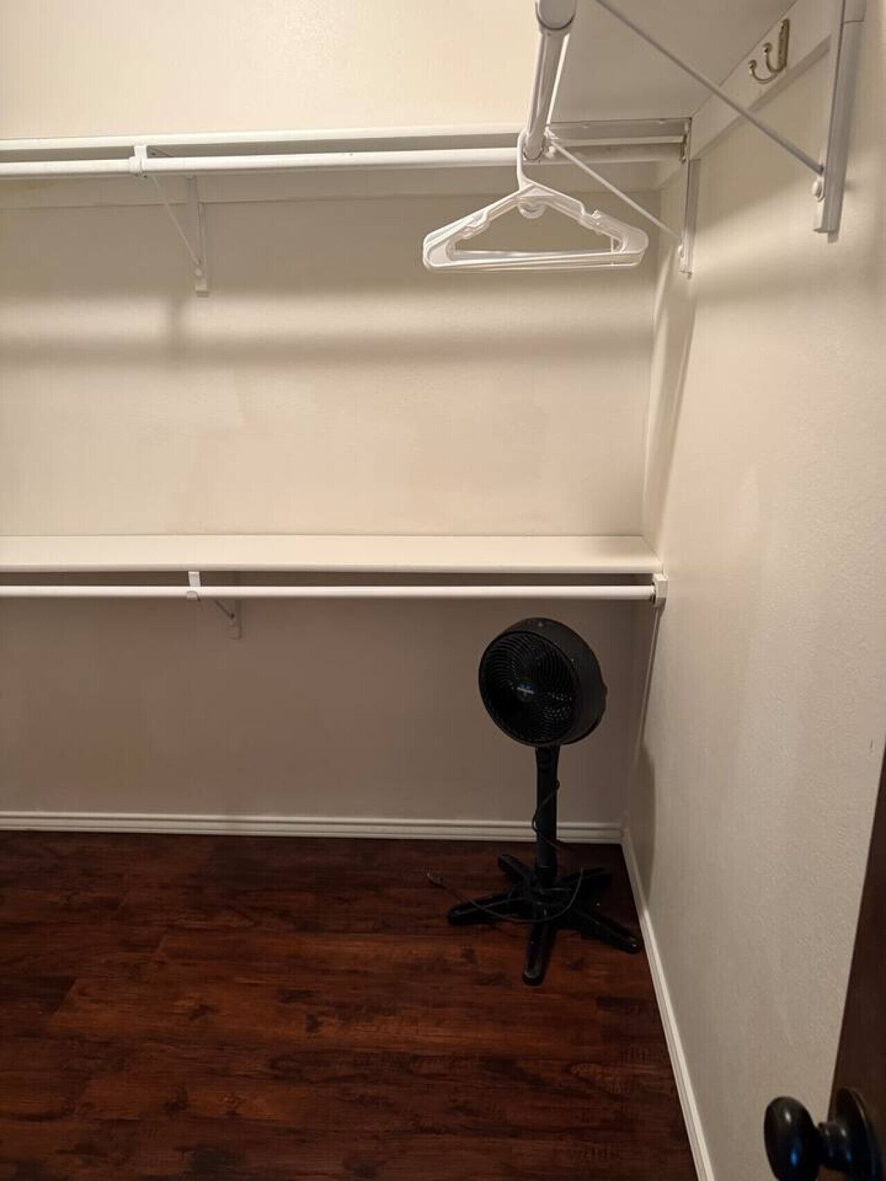 A room with a wooden floor, a black standing fan, and a white wall with a hanging clothes rack.