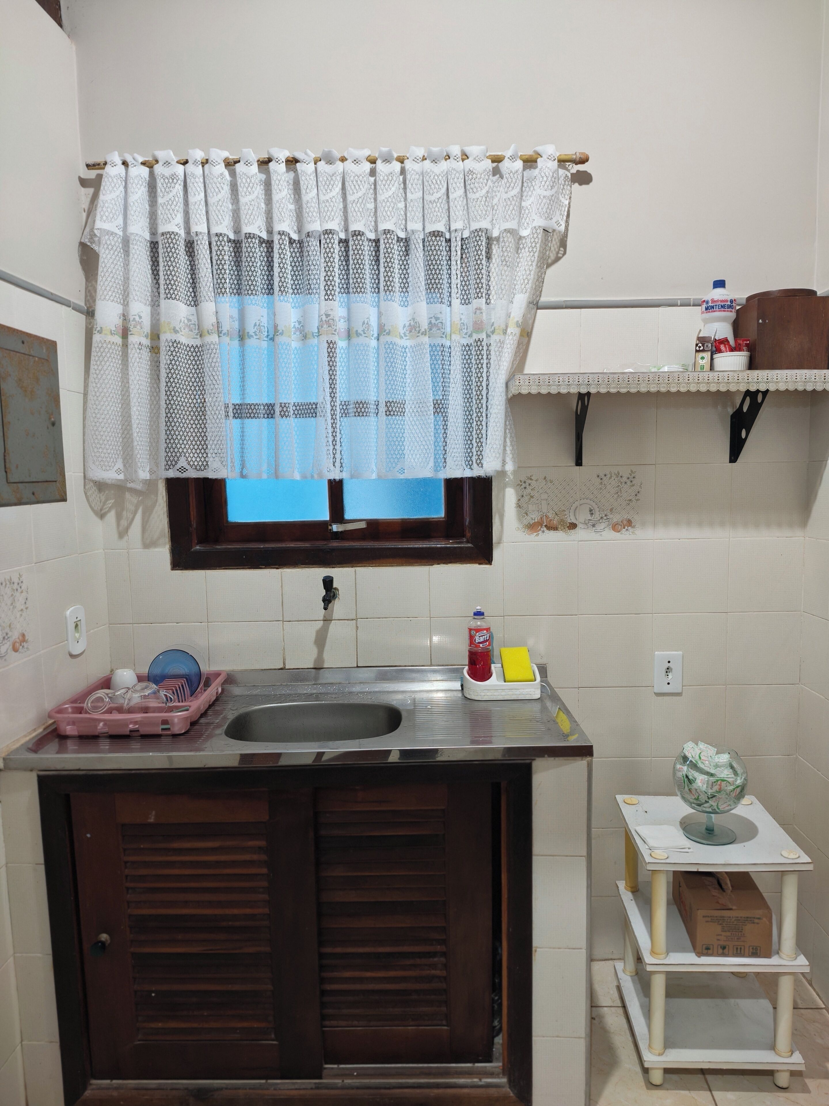 Una cocina compacta con un lavamanos, una ventana con cortina estampada, un estante pequeño con utensilios de cocina y una puerta.