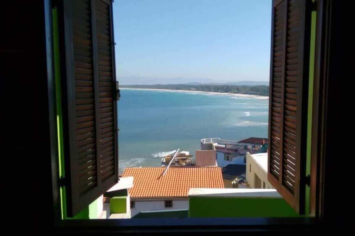 Vista costera a través de contraventanas abiertas, edificios con tejados de tejas rojas y un mar azul y despejado.