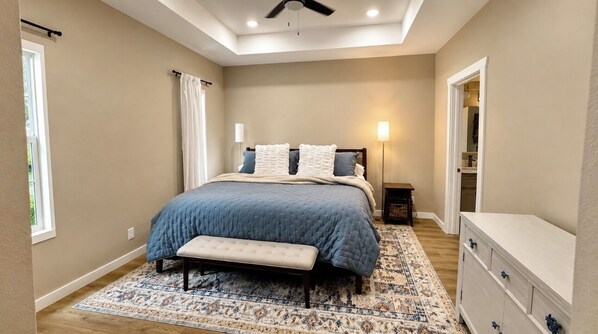 Quarto com cama, mesas de cabeceira, um banquinho, cômoda, ventilador de teto e tapete estampado.