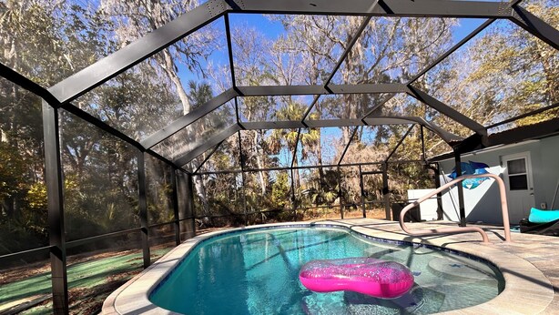 A swimming pool with a brick edge, surrounded by a glass enclosure with a metal frame, and a pink inflatable float. - 2-bedroom house w/pool in the heart of old Homosassa! (Homosassa)