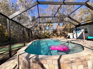 A swimming pool with a brick edge, surrounded by a glass enclosure with a metal frame, and a pink inflatable float. - 2-bedroom house w/pool in the heart of old Homosassa! (Homosassa)