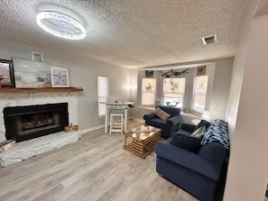 A living room with a fireplace, a blue sofa, a wooden coffee table, and a ceiling light fixture. - 2-bedroom house w/pool in the heart of old Homosassa! (Homosassa)