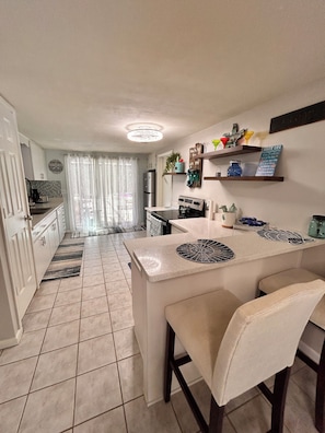 A modern kitchen with a central island, a refrigerator, and a dining area with chairs. - 2-bedroom house w/pool in the heart of old Homosassa! (Homosassa)