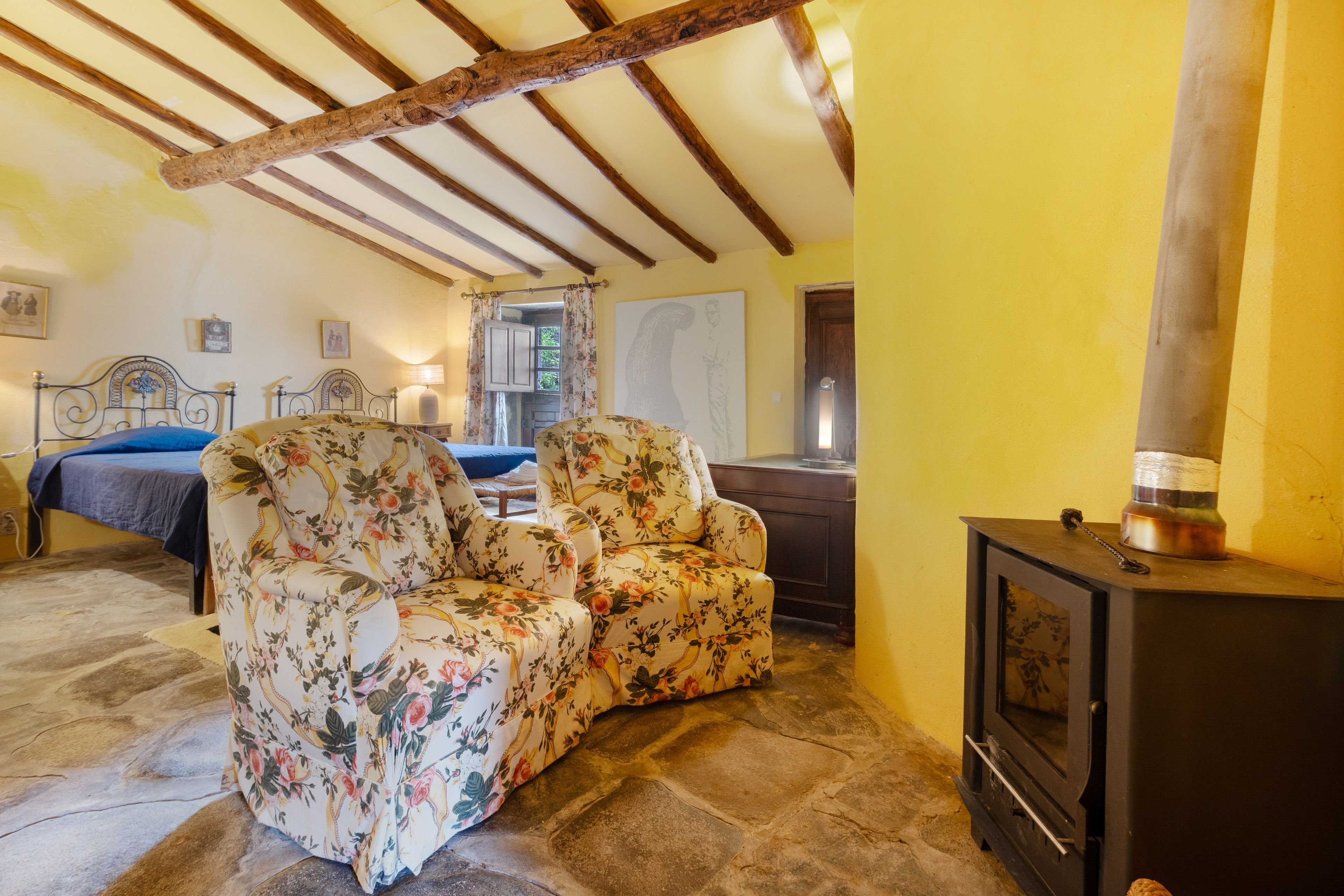 Una habitación con una cama, dos sillones florales, una mesa de madera y una chimenea.