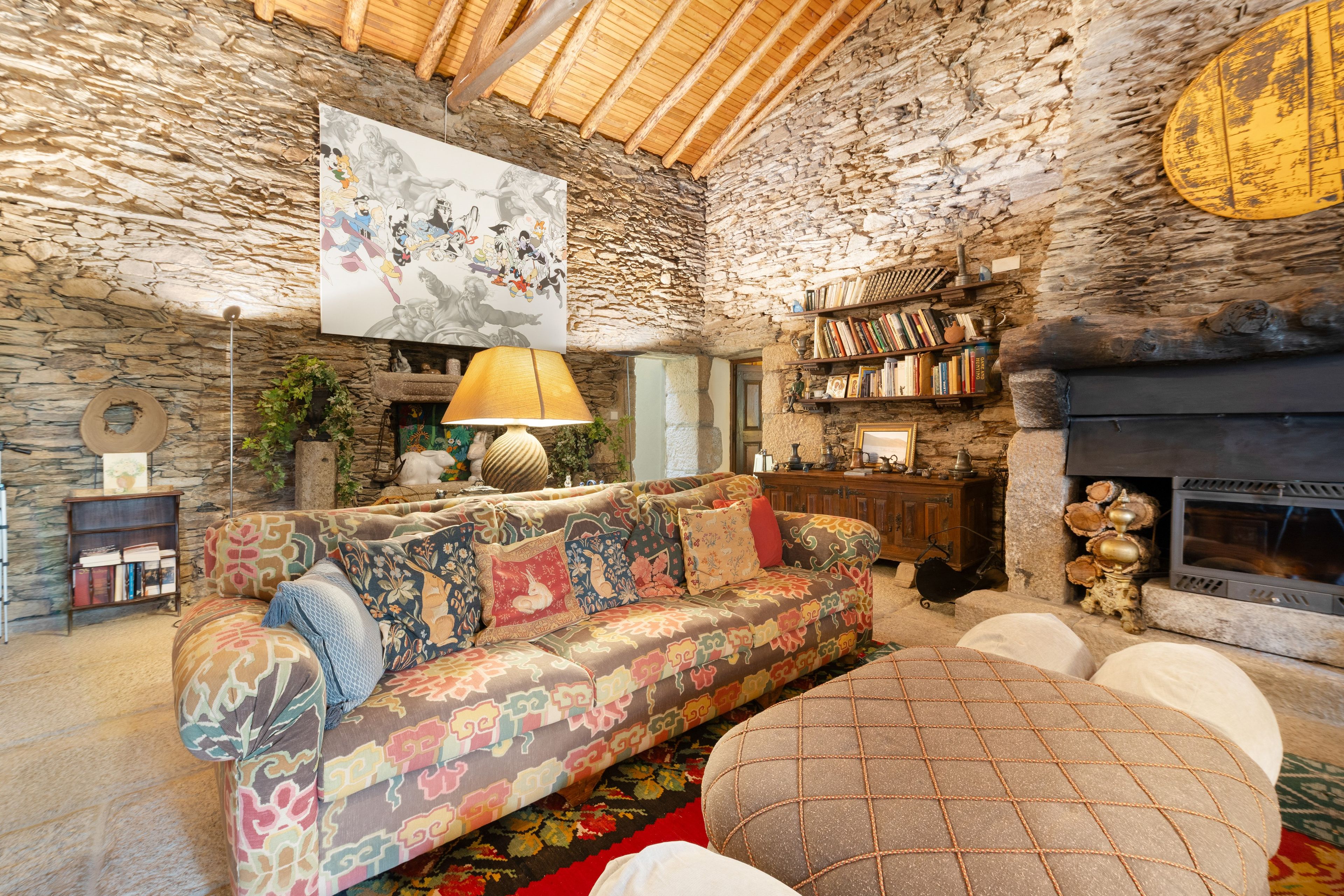 A cozy living room with a stone wall, a colorful sofa, a fireplace, and a bookshelf.