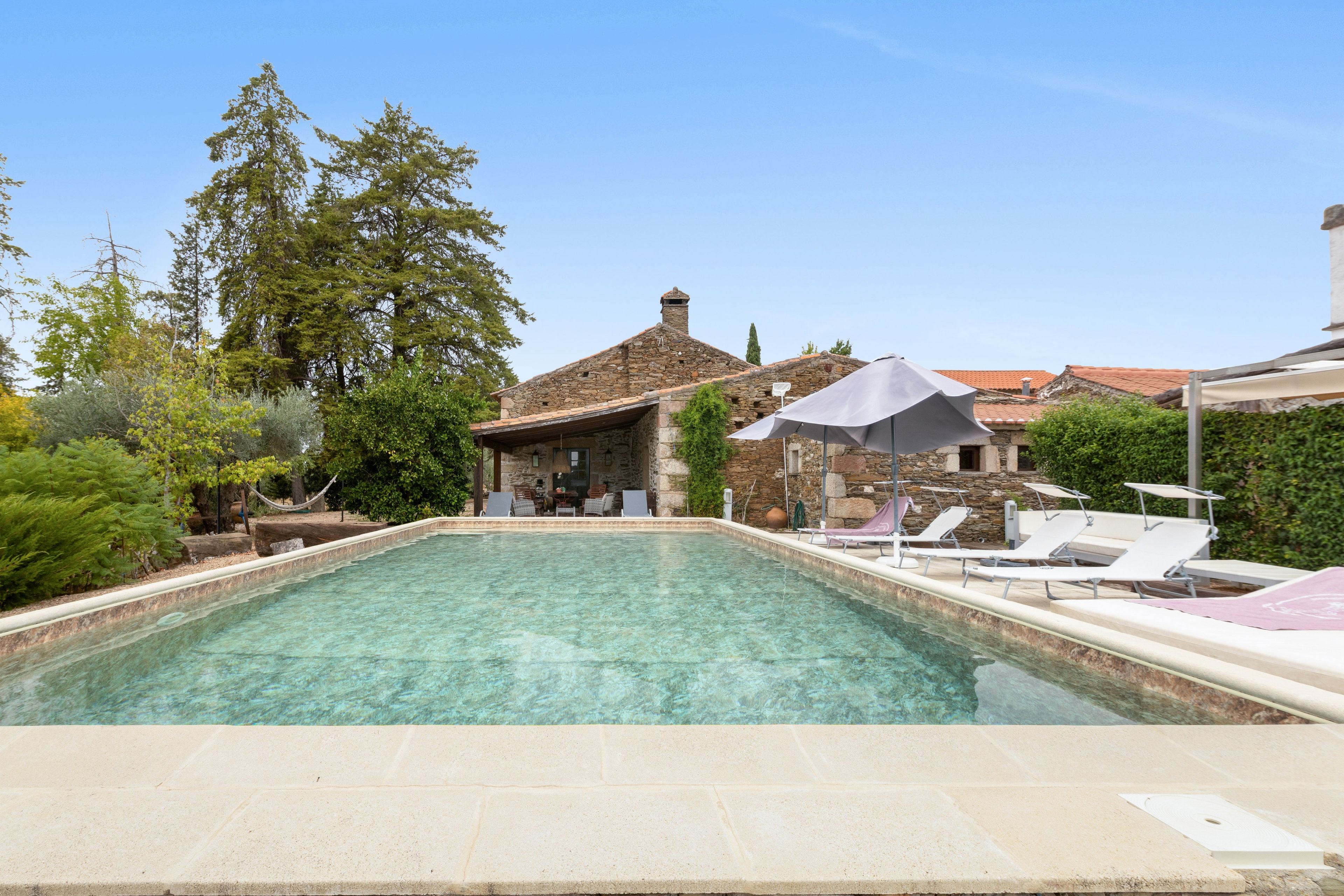 Una piscina con sillones de descanso y sombrillas, rodeada de árboles y un edificio de piedra.