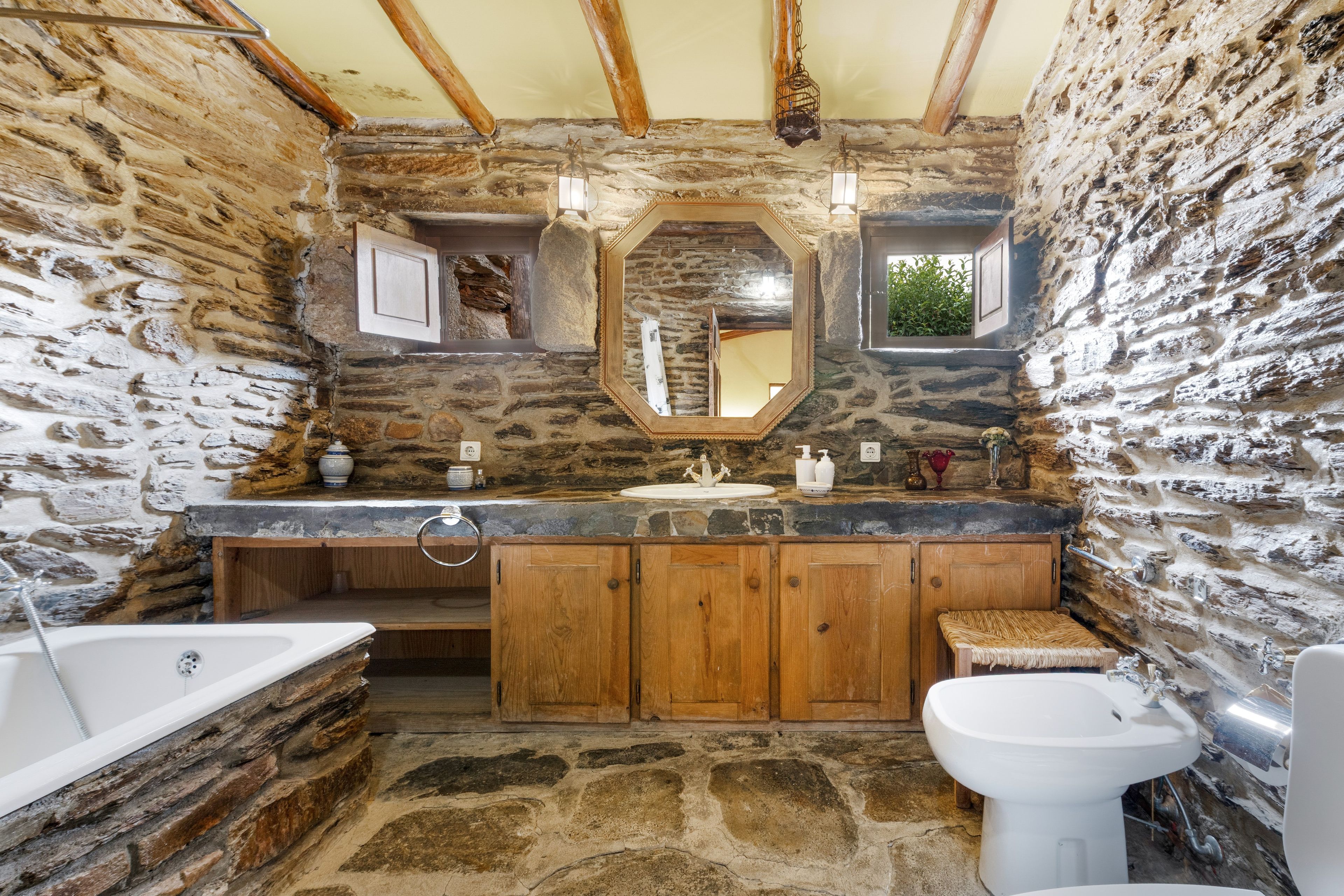 A rustic bathroom with stone walls, wooden cabinets, a stone sink, and a bathtub.