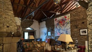 Un interior rústico con paredes de piedra vista, vigas de madera, un sofá estampado, una mesa de madera con diversos objetos y un cuadro abstracto de gran tamaño.