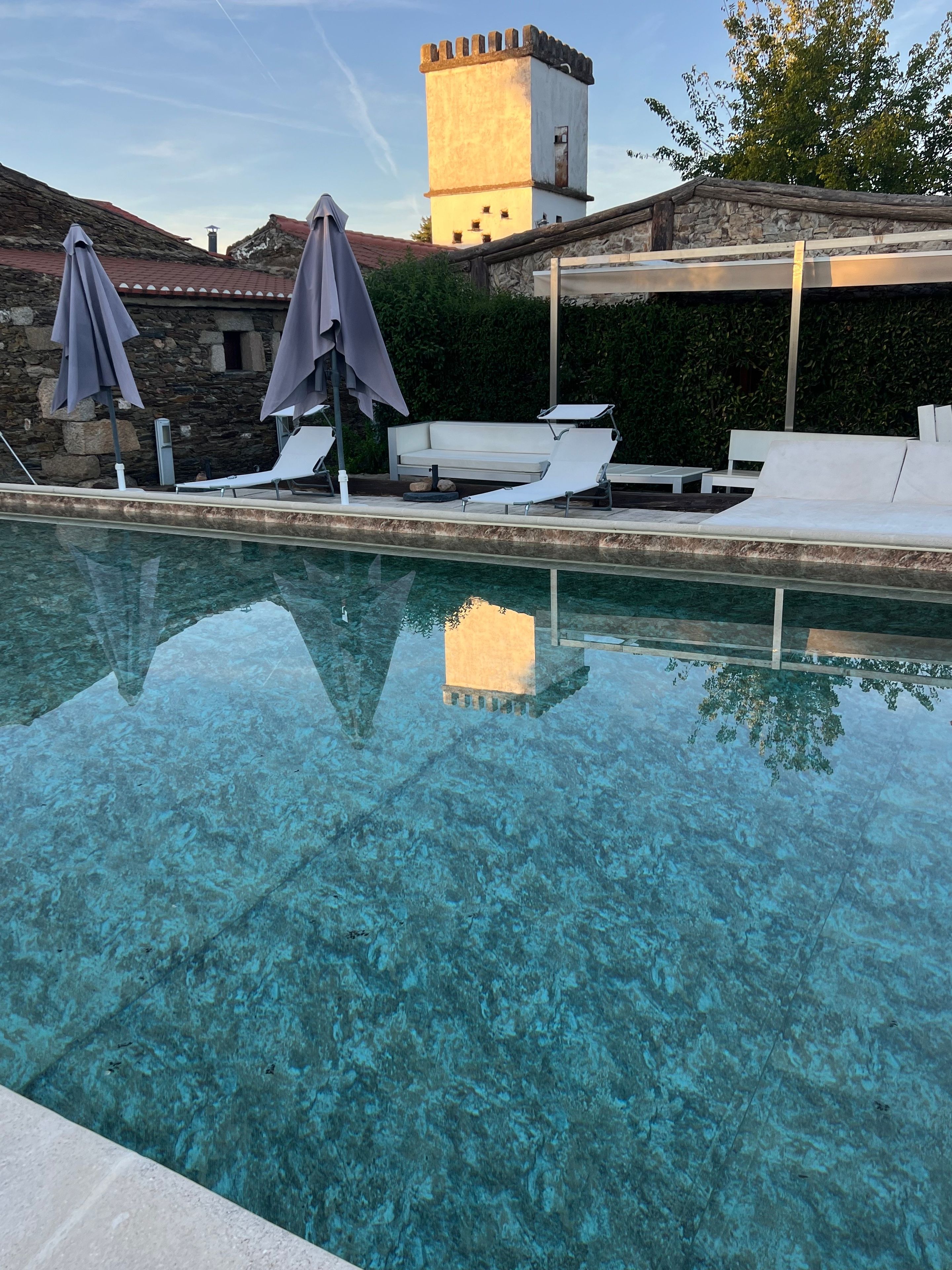 Une piscine aux eaux cristallines, dotée de chaises longues et de parasols, un mur de pierre et une tour en arrière-plan.