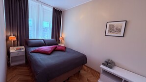 Un dormitorio con cama, dos mesitas de noche, una mesilla, una ventana con cortinas, un cuadro enmarcado en la pared y suelo de madera.