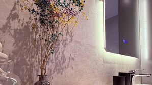 Un baño moderno con suelo de madera, un florero de vidrio con flores secas, un taburete pequeño y un espejo.