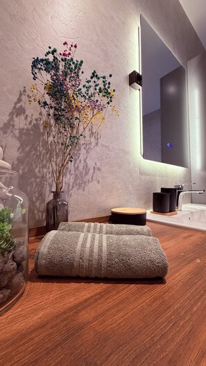 Ein modernes Badezimmer mit Holzboden, einer Glasvase mit getrockneten Blumen, einem kleinen Hocker und einem Spiegel.