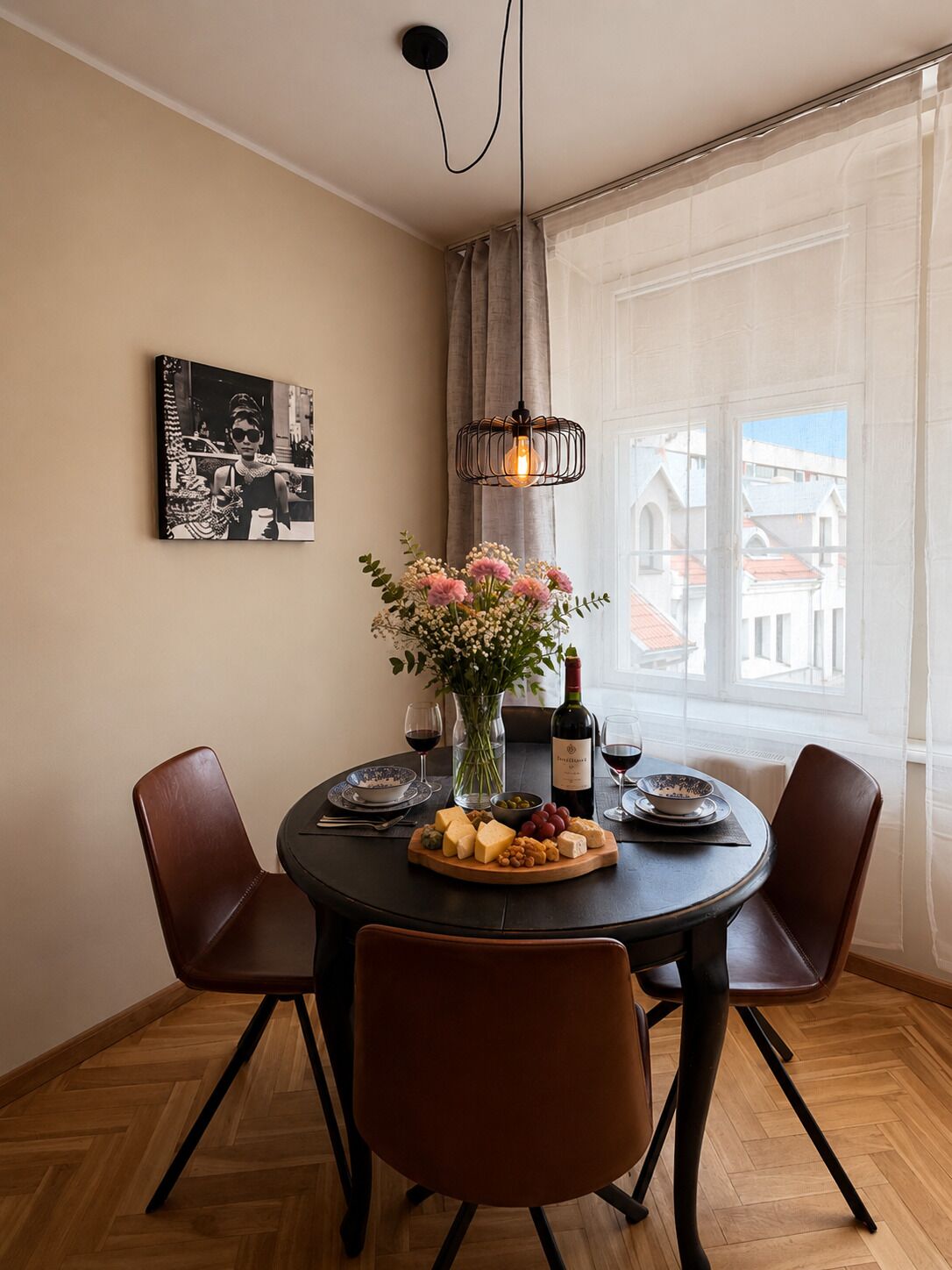 Un comedor con una mesa redonda, sillas, un jarrón con flores, una botella de vino y una fotografía blanco y negro en la pared.