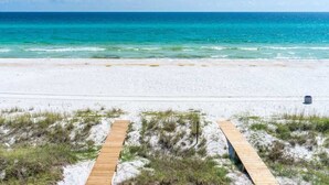 En strand med en träpromenad som leder ner till vattnet, en sandstrand och ett klart, blått hav.