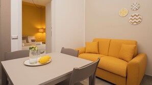 Una sala de estar moderna con un sofá amarillo, una mesa de comedor con una botella y un limón, y un pasillo que conduce a un dormitorio.