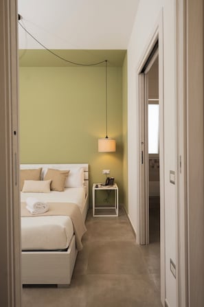 Ein schmaler Flur, der in ein Schlafzimmer mit Bett, Nachttisch, Lampe und einer Tür zu einem weiteren Raum führt.