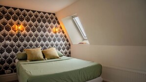 Une chambre avec un papier peint à motifs, un lit avec une literie verte, deux lampes fixées au mur et une lucarne.
