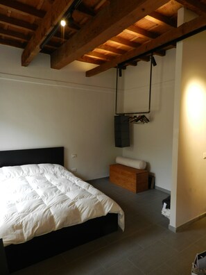 Ein Schlafzimmer mit Bett, Holzbalken, einer hängenden Leuchte, einer kleinen Holzbank und einem an der Wand befestigten Regal.