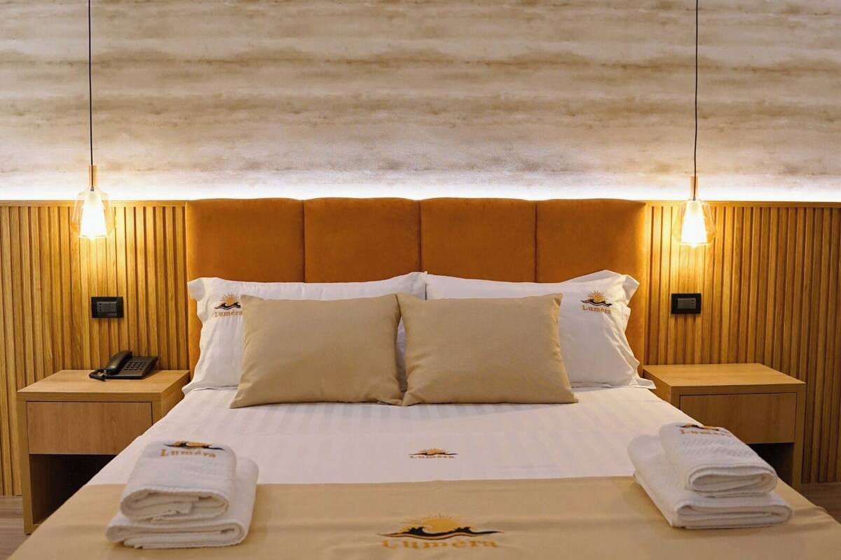 A hotel room with a wooden headboard, a bed with white linens, two bedside tables with lamps, and a telephone on one table.