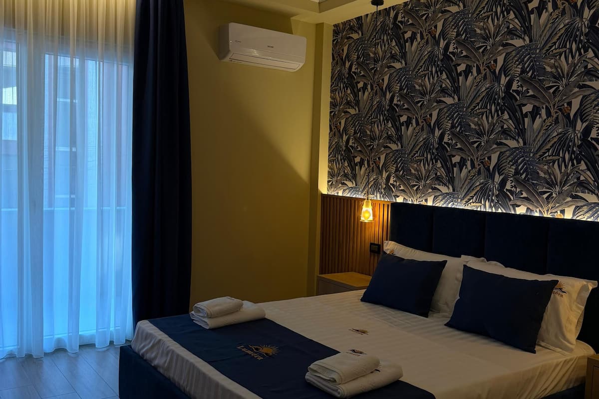 A hotel room with a bed, a nightstand, a wall-mounted air conditioner, and a window with curtains.