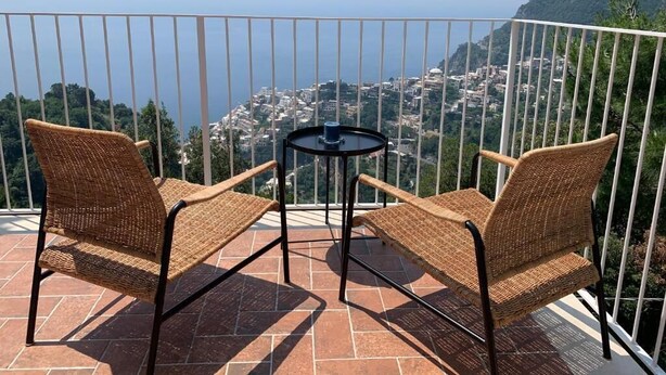 Un balcon avec des fauteuils en osier et une petite table, d’où l’on peut admirer un panorama urbain avec ses bâtiments et sa verdure.