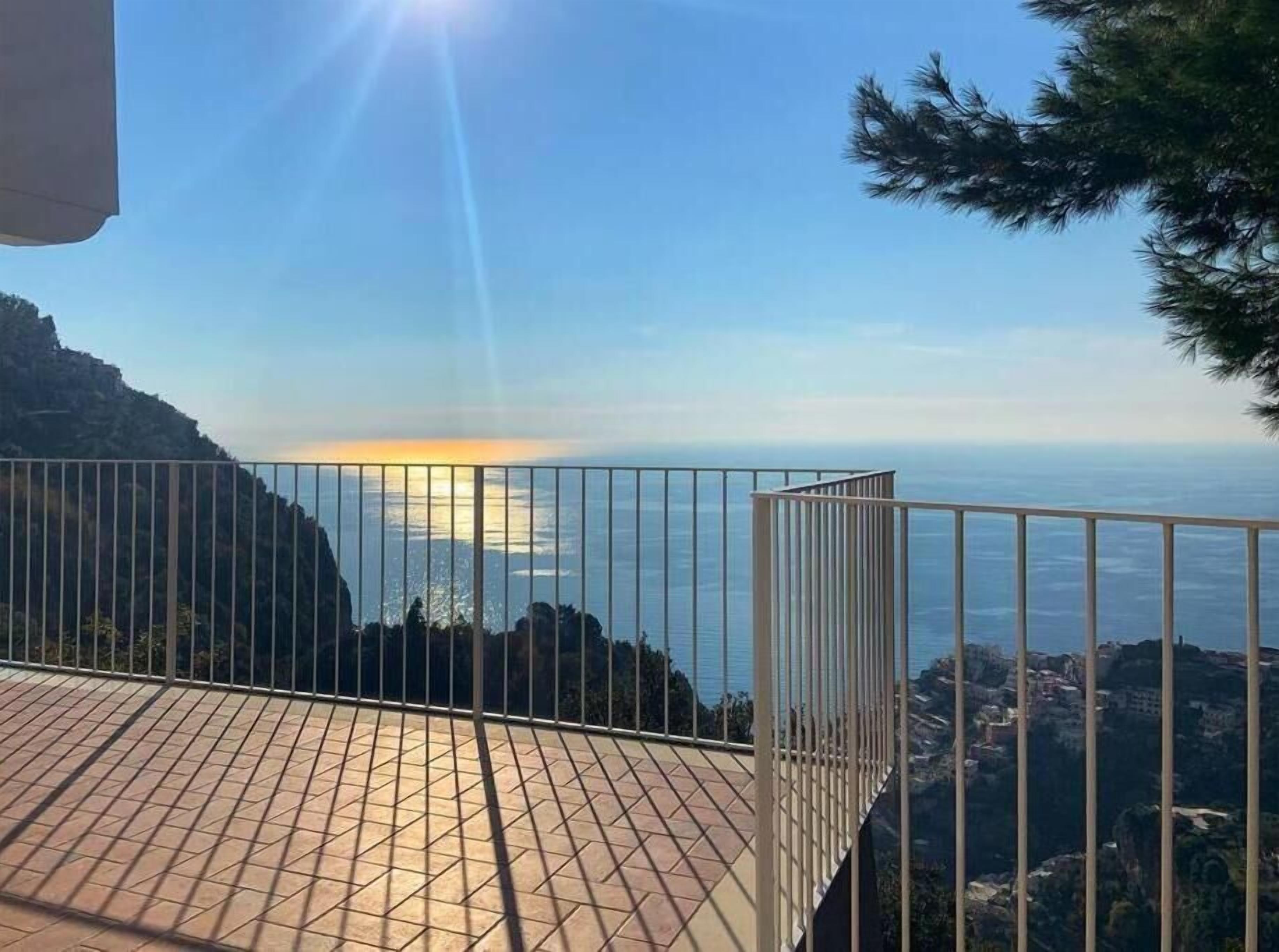 Un balcon doté d’un sol carrelé et d’une rambarde en métal, offrant une vue sur un paysage montagneux et la mer.