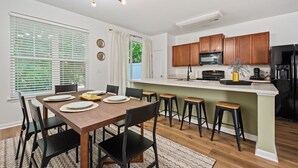 A modern kitchen with wooden cabinets, a black refrigerator, and a dining area with a wooden table and black chairs. - Rocking Oak Cottage of Charleston (Summerville)
