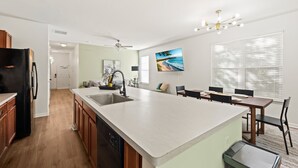 A modern kitchen with a large island, a dining area with a table and chairs, and a living room with a sofa and a TV. - Rocking Oak Cottage of Charleston (Summerville)