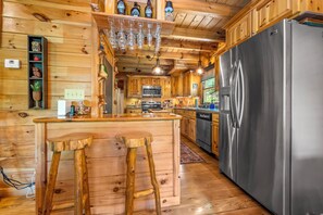 A wooden cabin interior with a kitchen featuring a stainless steel refrigerator, wooden bar stools, and a hanging light fixture. - Salty Pines- Guest Favorite (Lake Lure)