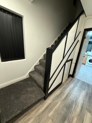 A staircase with carpeted steps and a black railing, leading to a room with a window and a view of a laundry area. - Rest and Recover (West Bromwich)
