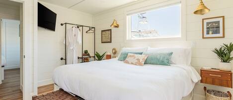 A bedroom with a large bed, a wooden bench, a bedside table with a plant, and a window with a view of a building.