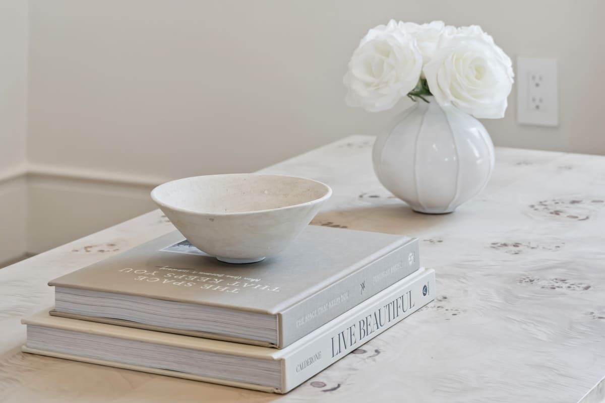 A white ceramic bowl on top of a stack of books, with a vase of white flowers to the right.