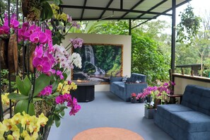 Un espace extérieur aménagé, abrité et doté d’un canapé et de fauteuils, entouré d’orchidées en pot et agrémenté d’un tableau représentant une cascade.