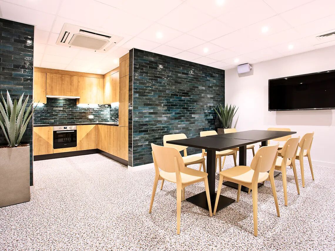 A modern kitchen with a built-in stove, a dining area with a black table and beige chairs, and a flat-screen TV mounted on the wall.