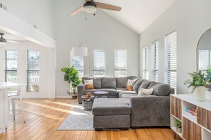 Ein geräumiges Wohnzimmer mit Ecksofa, Deckenventilator und großen Fenstern mit Jalousien.