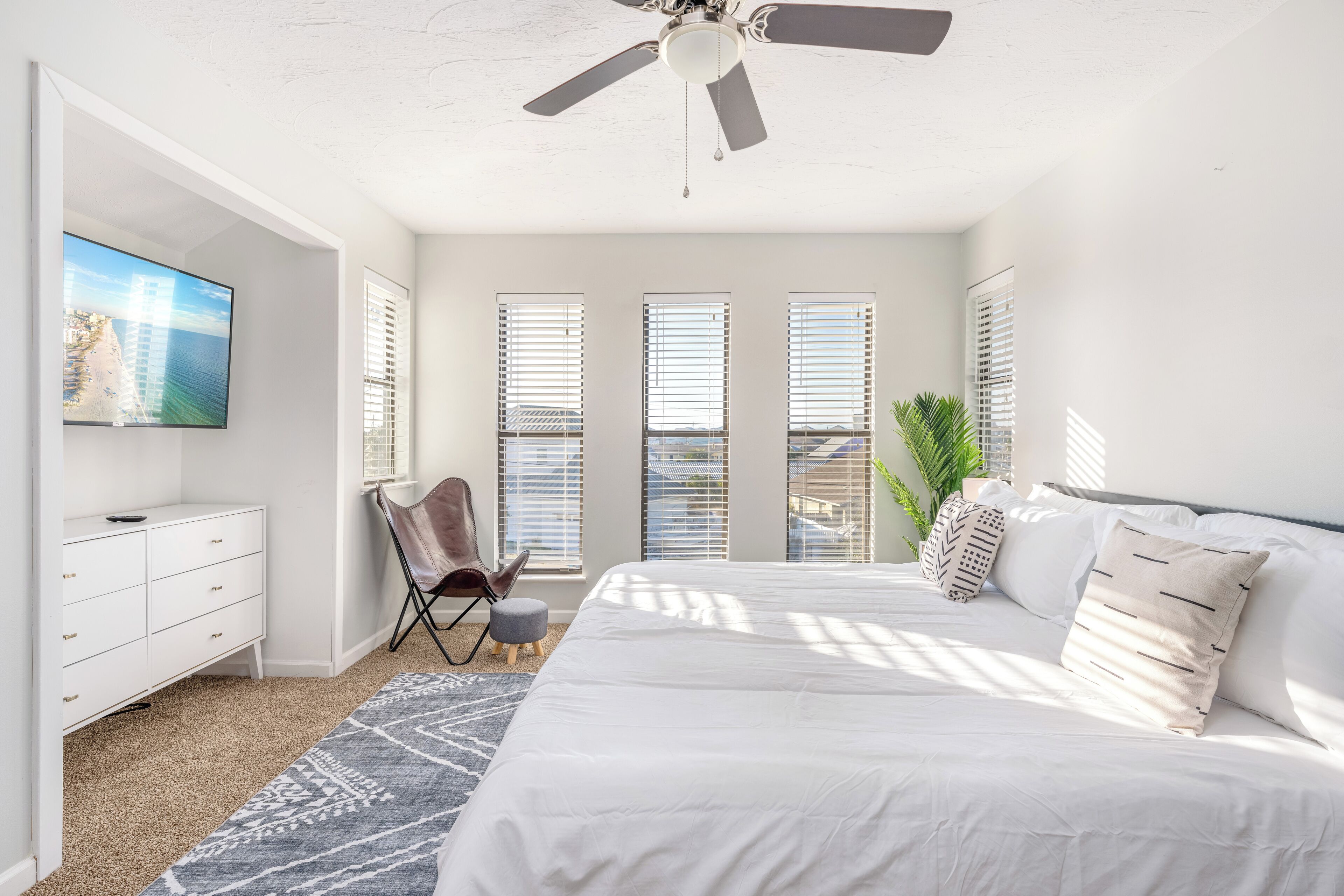 Un dormitorio con cama, ventilador de techo, televisor, escritorio, silla y ventanas amplias.