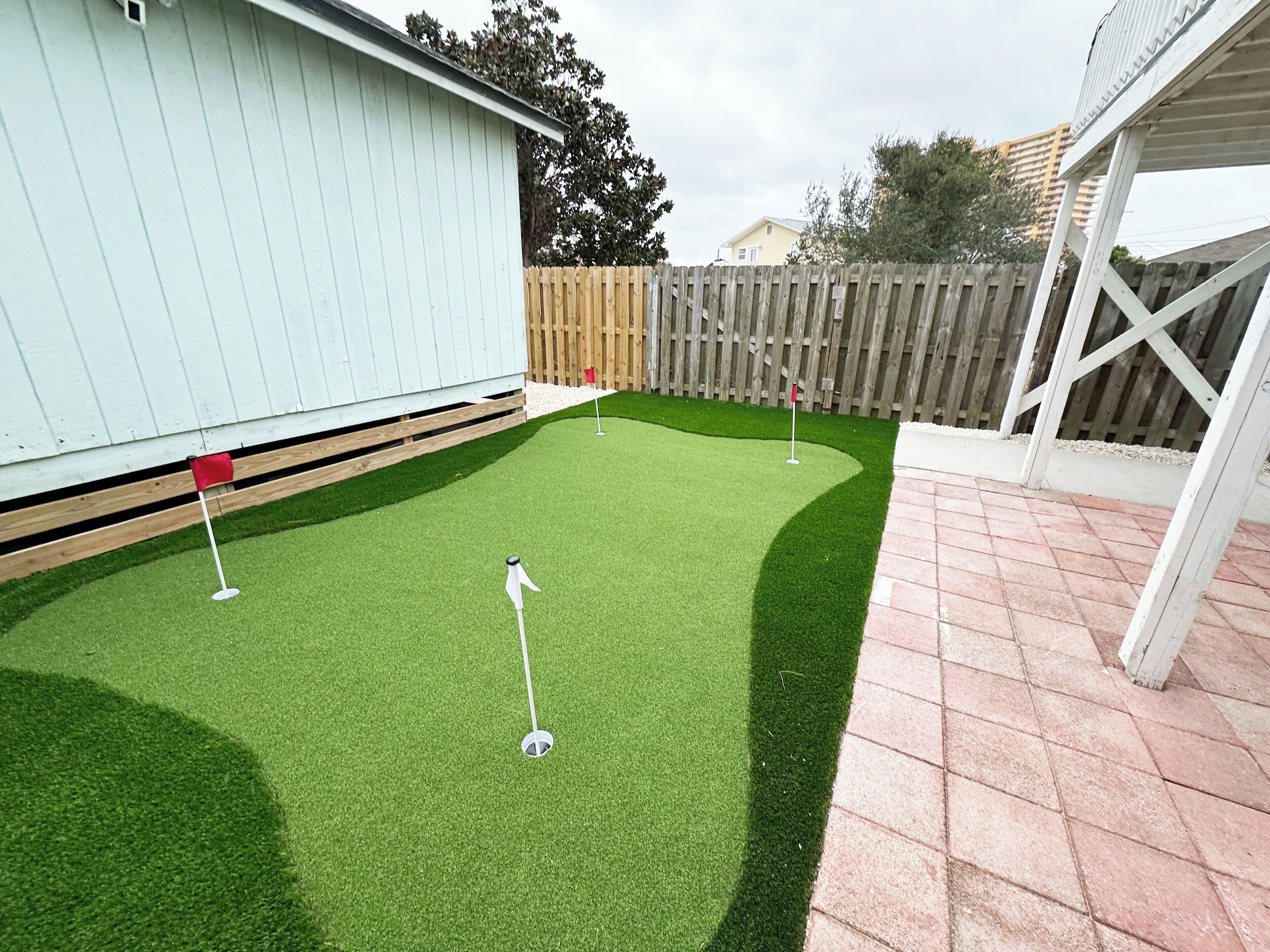 Un patio trasero con césped artificial verde, cerca de madera y un edificio de color azul claro.